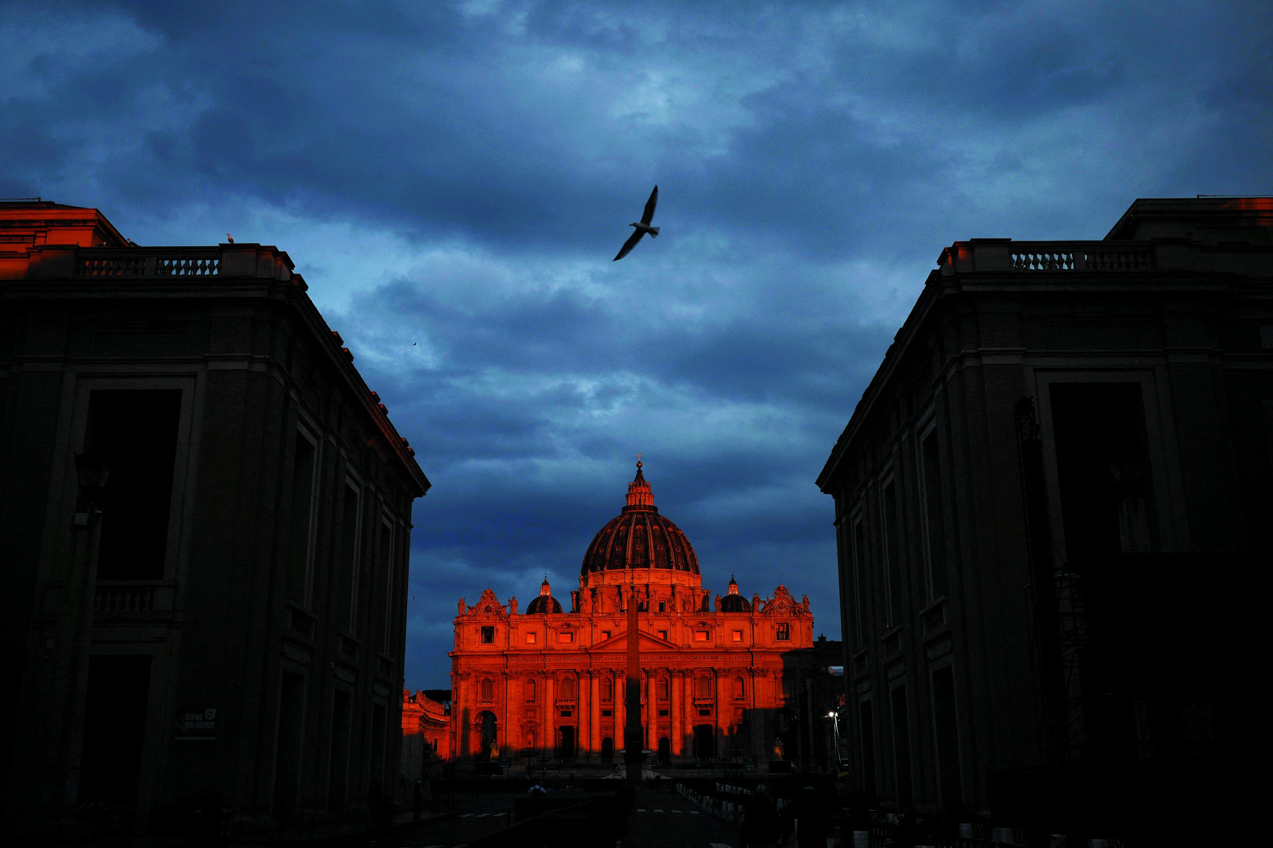 Vista da Basílica de São Pedro dias antes do conclave para eleger o novo papa, na Praça de São Pedro, no Vaticano visto de Roma, Itália, 28 de abril de 2025
Amanda Perobelli/REUTERS

Após a morte do Papa Francisco, em 21 de abril de 2025, a Igreja entrou em período de sede vacante. O funeral abriu um intervalo de cerca de onze dias até o início do Conclave. Nesse tempo suspenso, cardeais de diferentes partes do mundo chegaram a Roma para as congregações preparatórias, enquanto a Praça de São Pedro assumia outra função: não a do anúncio, mas a da espera. Entre colunas e pórticos, o cotidiano seguia em ritmo contido — freiras atravessavam o espaço, pequenos grupos se formavam e se desfaziam, olhares se voltavam para o interior de um processo que permanecia invisível.
O Conclave começou em 7 de maio de 2025. Ao todo, 133 cardeais — o maior número já reunido — entraram em isolamento na Capela Sistina, reiterando um ritual mantido por séculos, feito de gestos repetidos e decisões tomadas fora do alcance público. Foram necessárias quatro rodadas de votação para a escolha do novo pontífice: Robert Francis Prevost, 69 anos, que adotou o nome Leão XIV, em referência a Leão XIII.
Do lado de fora, a espera acumulada encontrou seu desfecho no anúncio do Habemus Papam. A praça, marcada dias antes pela contenção e pela observação silenciosa, reagiu de forma coletiva. Entre a solidez da arquitetura e a presença humano: o tempo. A fé vivida no intervalo e a tensão discreta entre permanência e mudança que atravessa a história da Igreja.